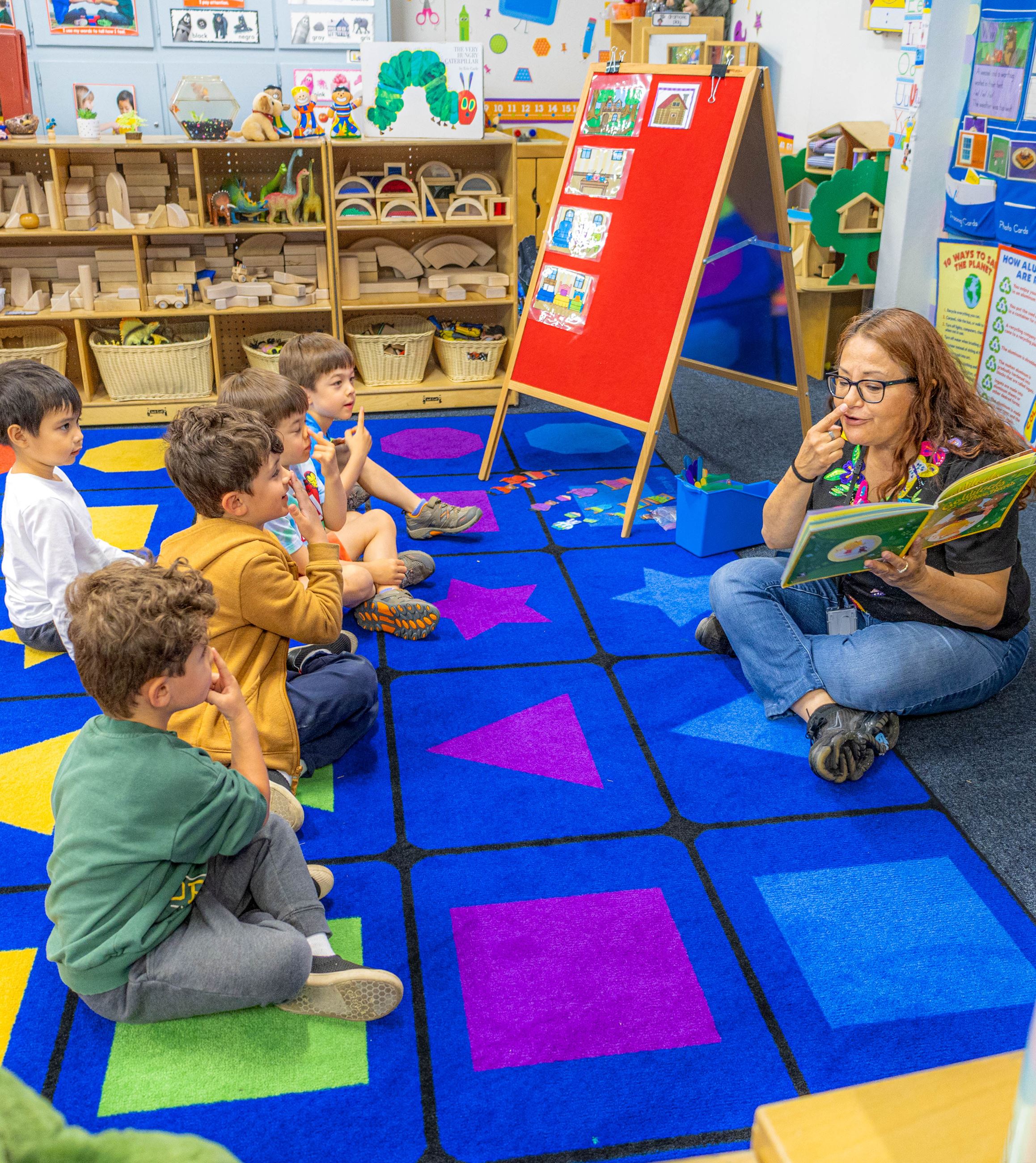 A preschool teacher reads a book to her students and touches her nose. Students mirror the action.