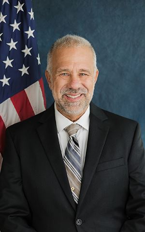 Portrait of Director Peter Sakaris sitting in front of the US Flag