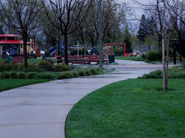 Gray cement walkway winding through the park past seating area, tennis courts and play area